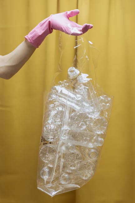 A person wearing a pink rubber glove is holding a clear plastic bag filled with numerous empty transparent plastic bottles, some crushed or flattened, which are typical recyclable waste. The bag appea