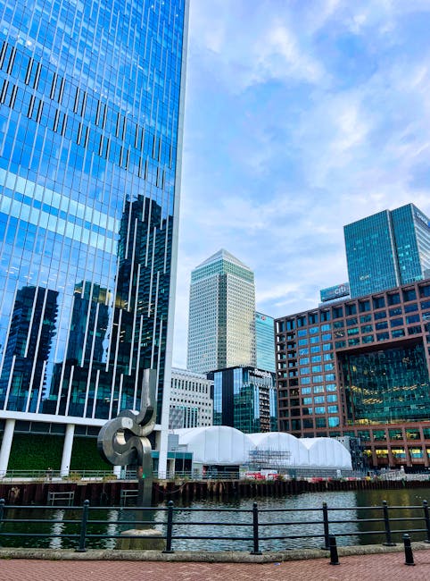 A modern urban scene featuring a red passenger train crossing a bridge over a river, with the train's exterior showing a sleek, metallic finish and vibrant red colouring. Behind the bridge, there are