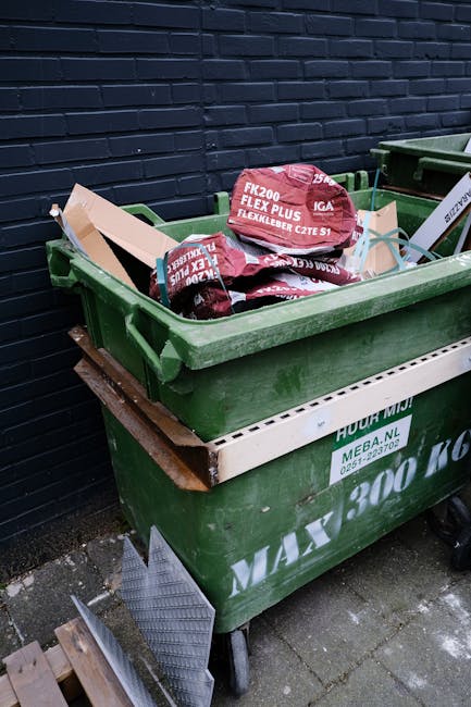 Two large black plastic rubbish bags, appearing scrunched and slightly torn, sit on the pavement near the curb in front of a dark wooden fence with vertical slats and a lattice panel on the left. The